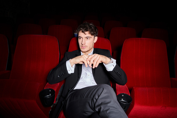 bello cercando elegante modello maschile con stile elegante seduto su sedie cinema rosso e guardando altrove - Foto, immagini