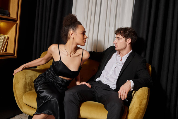 african american woman in black dress looking at elegant man in suit and sitting on velvet sofa - Photo, Image