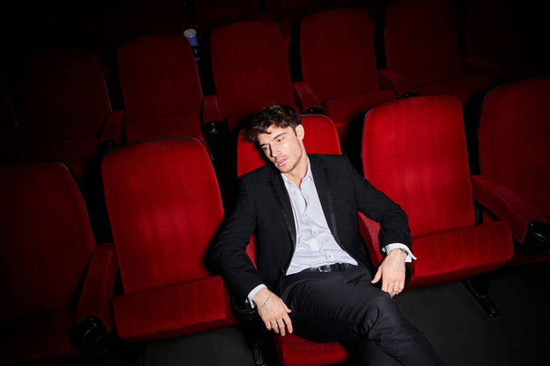 handsome elegant male model in black stylish suit sitting on red cinema chair and looking away - Zdjęcie, obraz