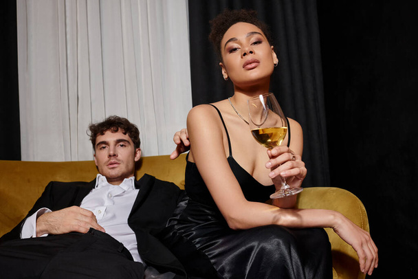 giovane donna afroamericana che tiene un bicchiere di vino e siede con l'uomo in giacca e cravatta sul divano di velluto - Foto, immagini
