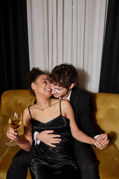 man hugging cheerful african american woman holding glass of wine and sitting on velvet sofa - Photo, Image