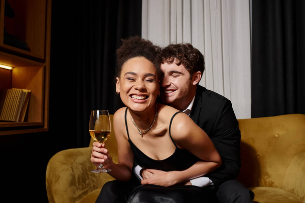 glücklicher Mann umarmt freudige afrikanisch-amerikanische Frau mit einem Glas Wein und sitzt auf dem Samtsofa - Foto, Bild
