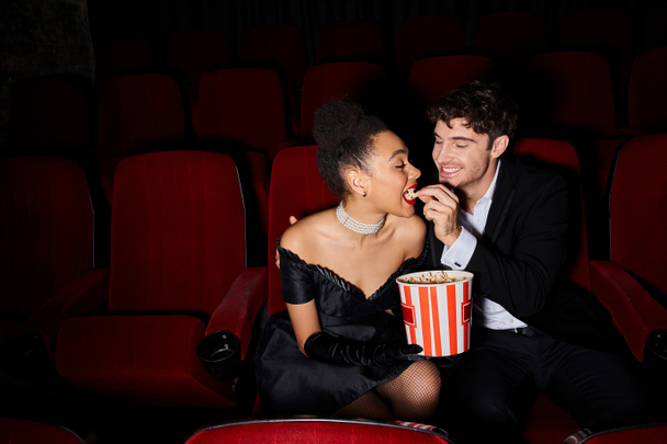 freudig schöner Mann im schwarzen Anzug füttert seine junge afrikanisch-amerikanische Freundin bei einem Date mit Popcorn - Foto, Bild
