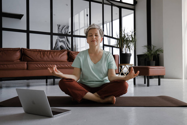 Женщина средних лет практикует йогу дома. Женщина с легкой улыбкой на лице сидит на коврике на полу в позе лотоса, использует ноутбук, чтобы смотреть расслабляющие видео и слушать медитативные - Фото, изображение