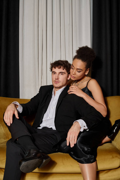 attractive african american woman in dress embracing man in formal wear and posing on velvet sofa - Photo, Image