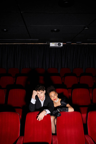 ελκυστικό πολυπολιτισμικό κομψό ζευγάρι σε μαύρο φόρεμα βράδυ κάθεται σε κόκκινες καρέκλες στον κινηματογράφο - Φωτογραφία, εικόνα