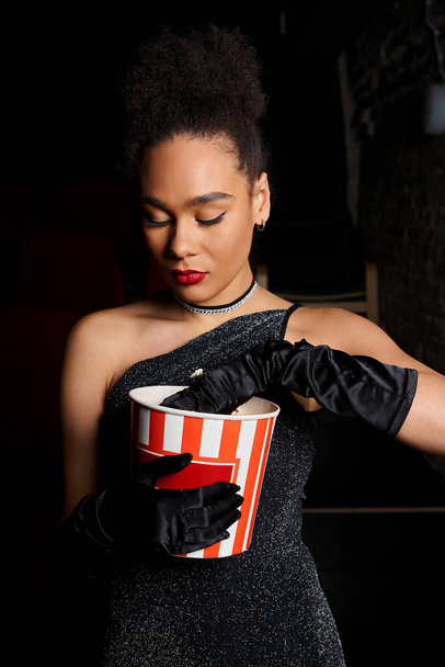 młody kuszące Afryki amerykanka kobieta w wieczór czarny sukienka z czerwony szminka pozowanie z popcorn - Zdjęcie, obraz