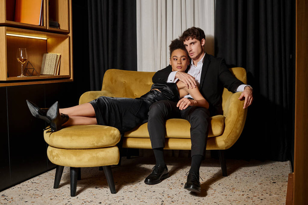 beautiful multicultural couple in black evening attire posing together while sitting on velvet sofa - Photo, Image