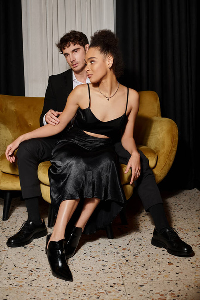 coppia multiculturale in abito da sera nero in posa insieme mentre seduti sul divano di velluto, eleganza - Foto, immagini