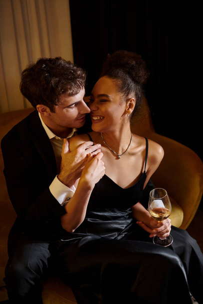 bell'uomo in giacca e cravatta che abbraccia felice donna afroamericana con bicchiere di vino e seduta sul divano - Foto, immagini