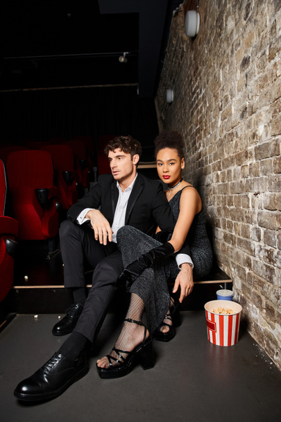 привлекательная многорасовая пара в черных элегантных одеждах, сидящих в кинотеатре на свидание в День Святого Валентина - Фото, изображение