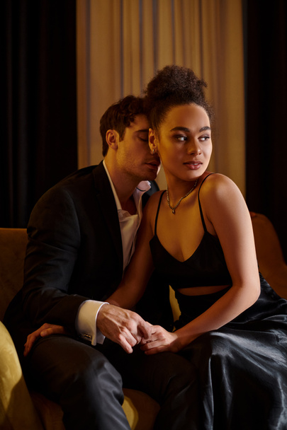 hombre guapo en traje seducir a mujer afroamericana bonita en vestido negro y sentado en el sofá - Foto, Imagen