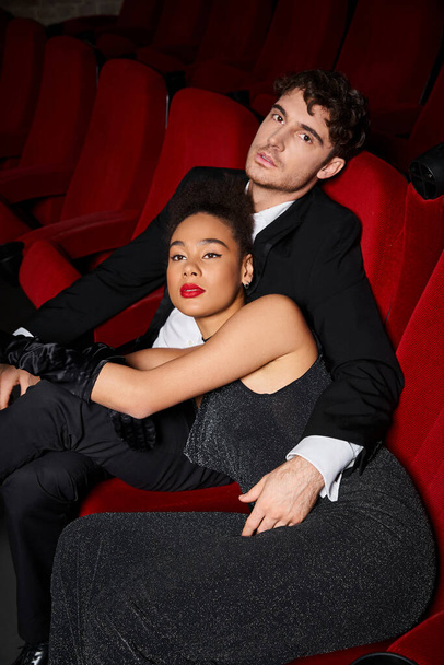attraktives multikulturelles Paar in schwarzen eleganten Kleidern posiert am Valentinstag liebevoll im Kino - Foto, Bild