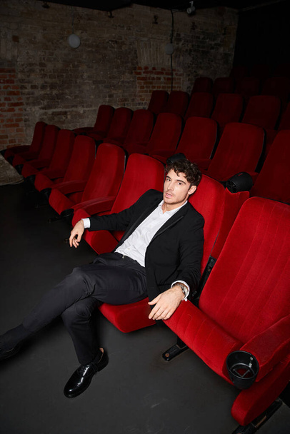 modelo masculino atractivo en elegante traje elegante sentado en la silla roja en el cine, mirando a la cámara - Foto, Imagen