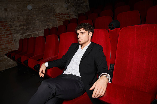 aantrekkelijk jong mannelijk model in stijlvol zwart pak zittend op rode stoel in de bioscoop en kijkend weg - Foto, afbeelding