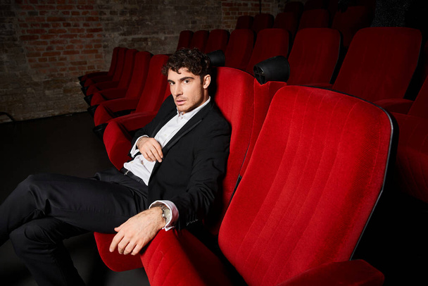 handsome appealing man in elegant black suit sitting on red chairs at cinema and looking at camera - Photo, Image