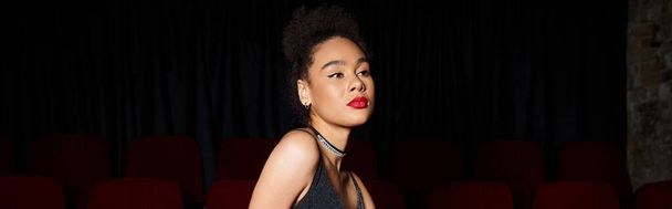 bela jovem afro-americana em vestido preto posando no cinema e olhando para longe, banner - Foto, Imagem