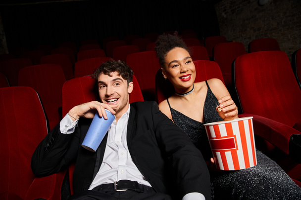 alegre pareja multiétnica en elegantes trajes disfrutando de la película en el cine, mientras que en la fecha, Día de San Valentín - Foto, Imagen