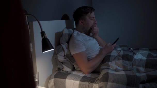 Homme caucasien souffre d'insomnie, ne dort pas la nuit, est accro à Internet et smartphone. Jeune homme se couche sur l'oreiller tard dans la nuit et regarde l'écran du téléphone mobile. Homme aux yeux fatigués. - Séquence, vidéo
