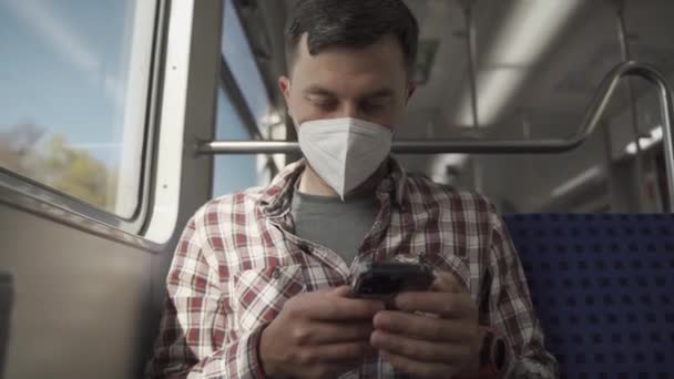 Uomo rilassato con maschera KN95 FFP2 sul viso utilizzando l'applicazione smartphone. Passeggero mascherato in viaggi ferroviari mentre seduto sul carro e sms sul suo cellulare. Viaggio sicuro sui trasporti pubblici.  - Filmati, video
