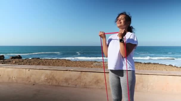 Mladé kudrnaté vlasy Latina žena v bílém tričku a šedé kamaše, cvičení s elastickými fitness pásky, cvičení na pažích, trénink její biceps svaly, stojící na pobřeží Atlantského oceánu - Záběry, video