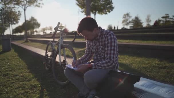 Estudante do sexo masculino tomando notas em um caderno sentado no parque perto de sua bicicleta. O tipo a estudar e a fazer os trabalhos de casa no parque a vir de bicicleta ao parque. Estilo de vida, educação ao ar livre. Campus universitário.  - Filmagem, Vídeo