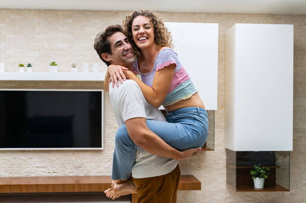 Side view of young cheerful couple in casual clothes hugging and standing at home while enjoying time together against TV with potted plants on shelves - Photo, image