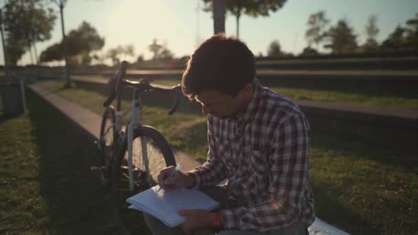 Student does his homework in the campus garden after a bicycle ride. Cyclist making notes in a sketchbook in the park. Learning in the outdoors. Healthy active lifestyle. Freelancer working outside.  - Footage, Video