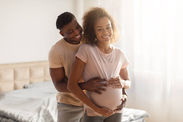 Молодая афроамериканская будущая пара разделяет любящий момент, муж нежно обнимает беременную жену, стоящую в современном интерьере спальни. Ожидание и радость будущего родительства - Фото, изображение