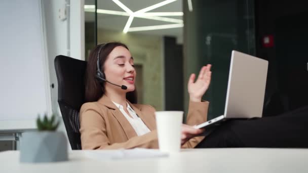 Beautiful female worker gesturing and leaning on back of chair during emotional video call with modern laptop in hands. Woman wearing wireless headset and business suit talking online at coffee break. - Footage, Video