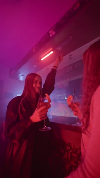 Video verticale, Due amiche danzano con cocktail nelle loro mani vicino al bancone del bar in un nightclub illuminato al neon. Donne felici Divertirsi a una festa in un bar notturno. Rallentatore. - Filmati, video