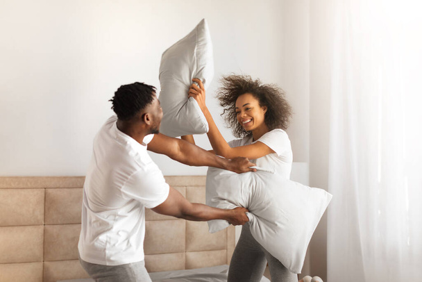 Radosna Afroamerykanska Para Tysiącletniego O Walce Poduszkowej Dla Zabawy W Sypialni W Domu. Młody mąż i żona walczą z poduszkami, śmieją się i flirtują w weekend. Szczęśliwego związku romantycznego - Zdjęcie, obraz