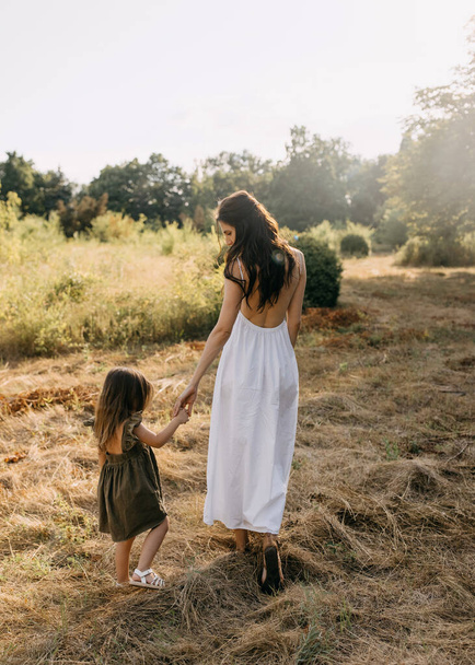 Moeder loopt en praat met haar dochter, hand in hand, in een veld op een zomerdag. Van achteren neergeschoten.. - Foto, afbeelding