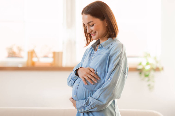 Hamile Avrupalı kadın karnına sevgi ve heyecanla dokunuyor ve sarılıyor, bebeğinin gelişini bekliyor, modern iç mekanının penceresinin yanında duruyor. Hamilelik, çocuk beklentisi - Fotoğraf, Görsel