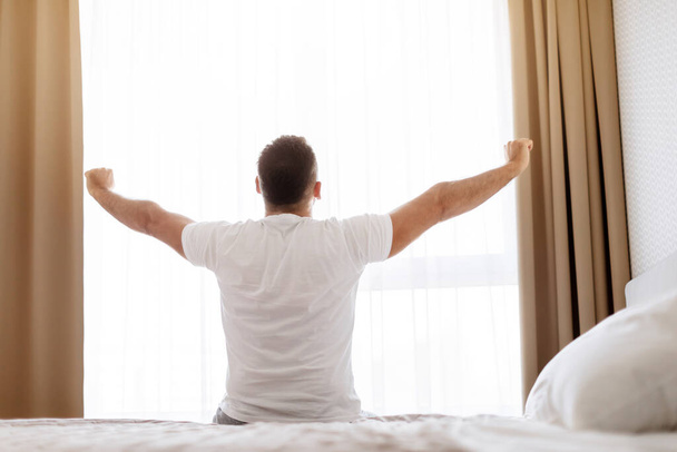 おはようございます。 ウィンドウを見ながら,モダンなベッドルームで快適なベッドに座って手を伸ばす男のリアビュー. 新しい日の始まり,休息とウェルネス - 写真・画像
