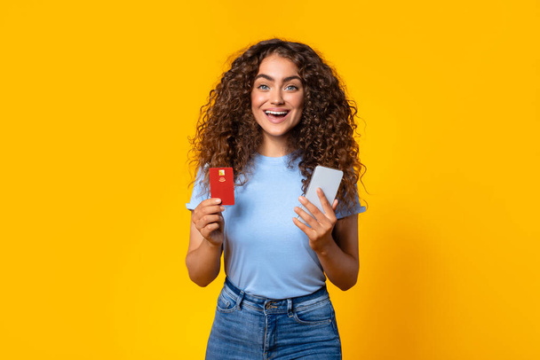 Donna estatica con i capelli ricci in possesso di una carta di credito e smartphone, pronto per lo shopping online, su uno sfondo giallo vivace - Foto, immagini