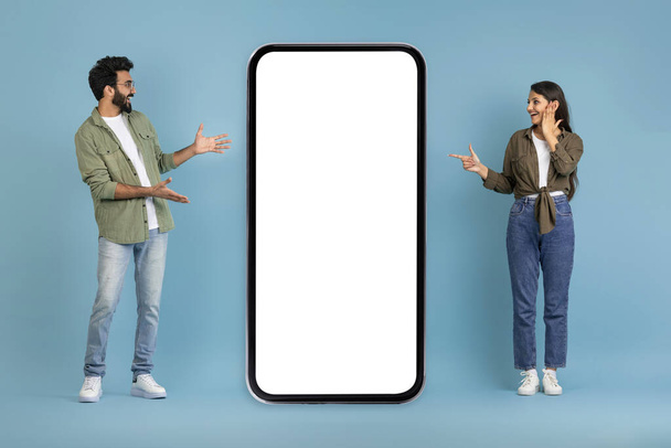 Amazed emotional millennial indian man and woman lovers pointing at big phone with white empty screen, showing great mobile app, online offer, blue studio background, mockup, blank space - Photo, Image