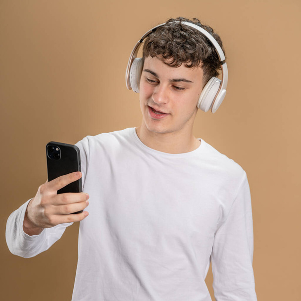 Jeden mężczyzna biały młody mężczyzna stoisko studio strzał na beżowym tle używać telefon komórkowy smartfon ze słuchawkami słuchać muzyki wysyłać wiadomości SMS lub przeglądać internet online app przestrzeń kopiowania - Zdjęcie, obraz