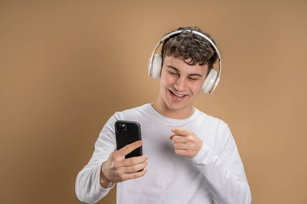 Ένας άνδρας καυκάσιος νεαρός άνδρας stand studio shot σε μπεζ φόντο χρησιμοποιούν κινητό τηλέφωνο smartphone με ακουστικά ακούσετε μουσική στείλετε μηνύματα γραπτών μηνυμάτων ή περιηγηθείτε στο διαδίκτυο online app αντίγραφο χώρου - Φωτογραφία, εικόνα
