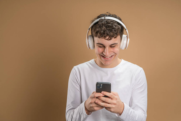Jeden mężczyzna biały młody mężczyzna stoisko studio strzał na beżowym tle używać telefon komórkowy smartfon ze słuchawkami słuchać muzyki wysyłać wiadomości SMS lub przeglądać internet online app przestrzeń kopiowania - Zdjęcie, obraz