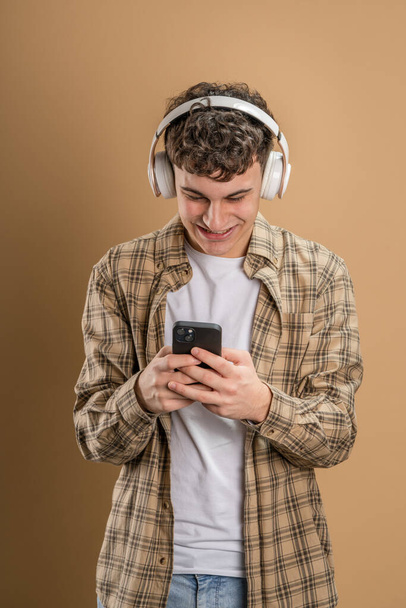 Beyaz tenli genç bir adam bej arka planda çekim yapıyor. Kulaklıklı cep telefonu kullanıyor. Müzik mesajlar gönderiyor ya da internetteki uygulamaları kopyalıyor. - Fotoğraf, Görsel