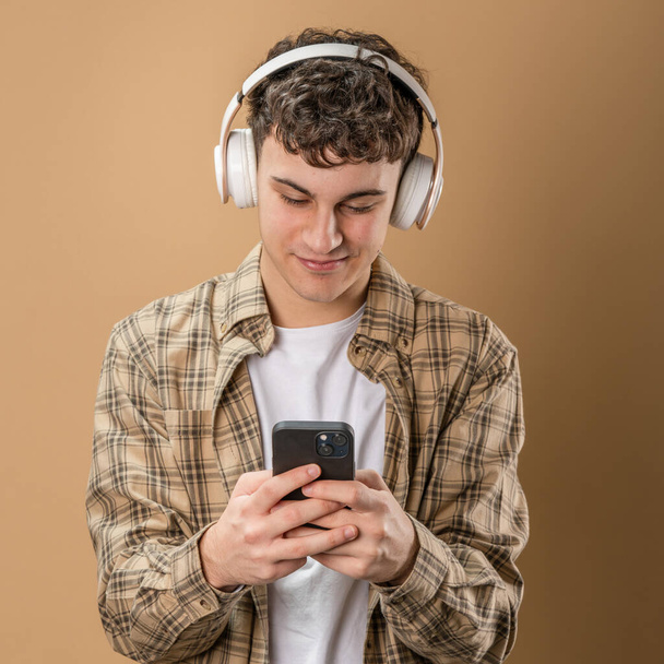 Yksi mies valkoihoinen nuori mies stand studio ammuttiin beige tausta käyttää matkapuhelimen älypuhelin kuulokkeet kuunnella musiikkia lähettää viestejä kutoma tai selata internetin online-sovellus kopioi tilaa - Valokuva, kuva