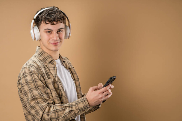 Один чоловік кавказький молодий чоловік стоїть студійний знімок на бежевому фоні використовуйте мобільний телефон з навушниками слухати музику надсилати смс-повідомлення або переглядати інтернет-простір копіювання додатків - Фото, зображення