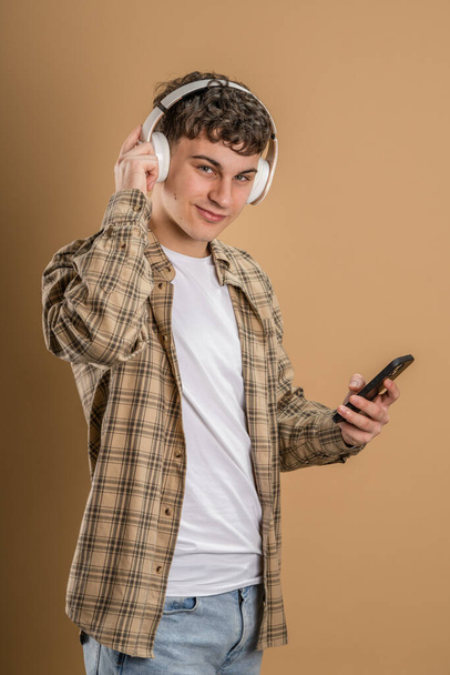 Один человек кавказский молодой мужчина стенд студии снят на бежевом фоне использовать мобильный телефон смартфон с наушниками слушать музыку отправить сообщения смс или просматривать интернет онлайн приложение копировать пространство - Фото, изображение