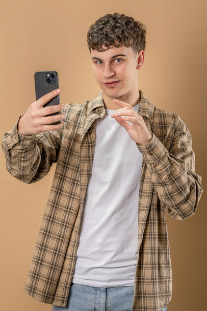One man adult caucasian male teenager stand studio shot happy smile use mobile phone smartphone to make a call talking video call or self portrait selfie copy space in front of beige background - Zdjęcie, obraz