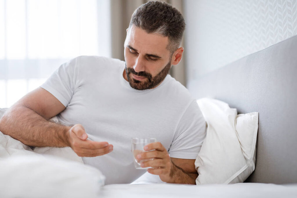 Sick Middle Aged Man Taking Medication Pill Holding Glass Of Water, Treating Fever Or Headache In The Morning, Lying In Bed In Modern Bedroom Interior. Sickness And Medical Treatment Concept - Foto, Bild