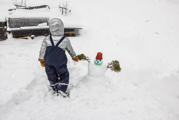 View of a child by a self-made cute snowman in a snowy garden. Sweden. - Photo, Image
