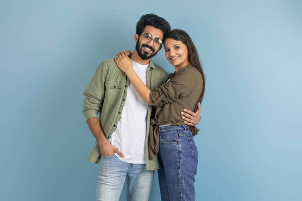 Gyönyörű Y-generációs indiai pár alkalmi ruhát visel, kék stúdió háttérben pózol, boldog fiatal keleti férfi és nő ölelkezik és mosolyog a kamerába. Szerelem, kapcsolatok, házasság - Fotó, kép