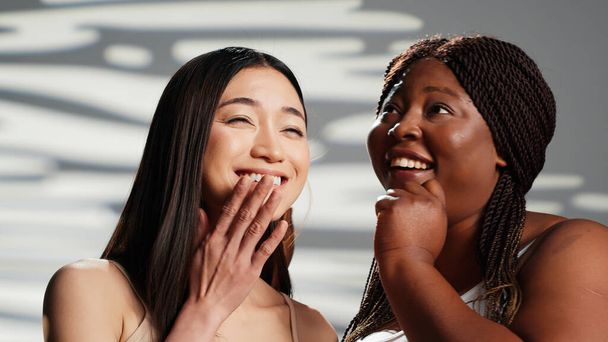 アフリカ系アメリカ人女性は、アジア系の女の子と秘密を共有し、スキンケアと自己愛キャンペーンを作成します。広告体の積極性と自己受容、異人種間の友人笑っている。美しい女の子. - 写真・画像