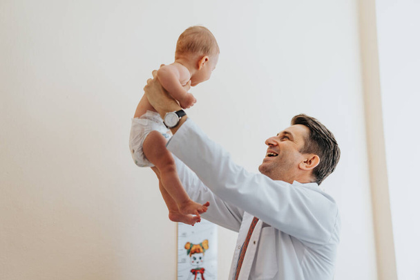 Щасливий чоловік, лікар-педіатр, який витягує маленьку дитину в клініці. Вони посміхаються. - Фото, зображення